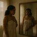 A woman (African, mid-to-late 20s or early 30s) in an off-white casual dress standing near a mirror, gazing at her reflection, unsure, wistful, a little broken.