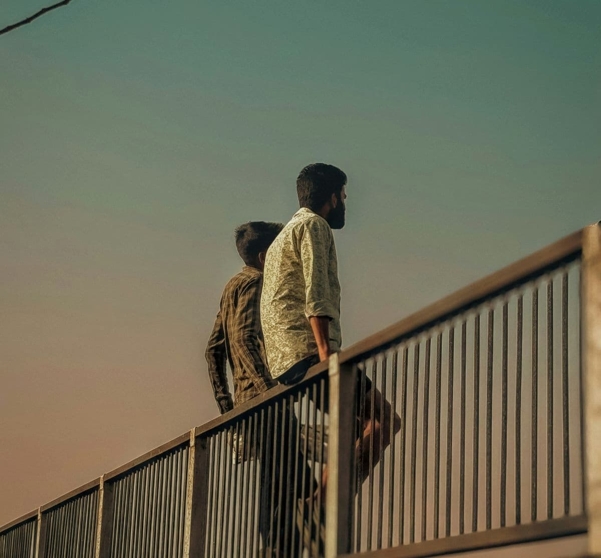 A back view photo of two men sitting on a rail with a small empty space beside them, evening light casting long shadows.