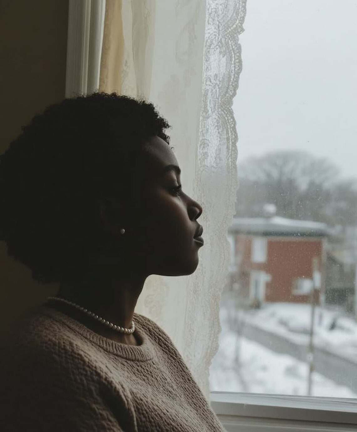 A young African woman sitting by a window at dawn with a mix of hope and sadness.