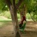 A woman wearing a red headscarf sits alone on a green bench in a park, partially obscured by a tree in the foreground.