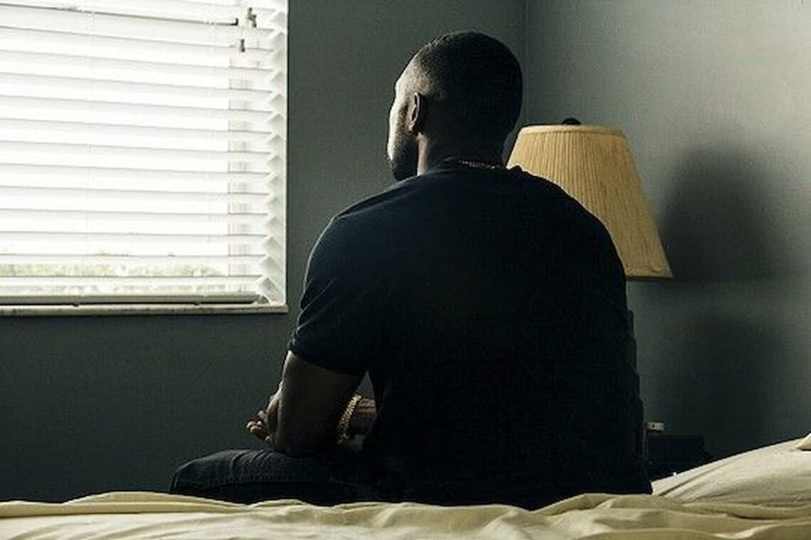 A young man sits alone on a bed in a dimly lit room, facing a window with closed blinds. He appears deep in thought, his hands clasped, as soft daylight filters in. A bedside lamp stands unlit beside him.