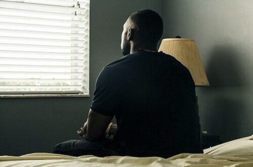 A young man sits alone on a bed in a dimly lit room, facing a window with closed blinds. He appears deep in thought, his hands clasped, as soft daylight filters in. A bedside lamp stands unlit beside him.
