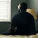 A young man sits alone on a bed in a dimly lit room, facing a window with closed blinds. He appears deep in thought, his hands clasped, as soft daylight filters in. A bedside lamp stands unlit beside him.