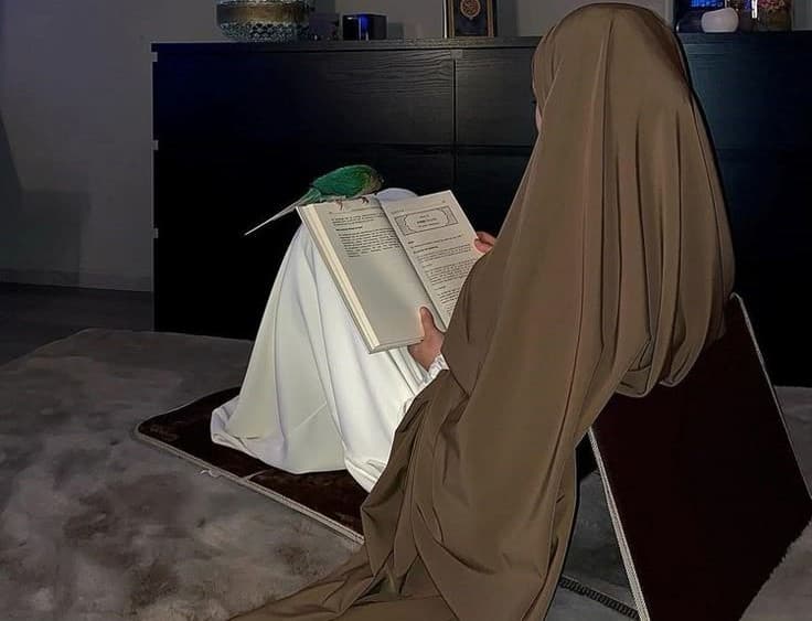 A Muslim woman in a simple hijab, seated on the floor of a modest room, reading the Quaran.