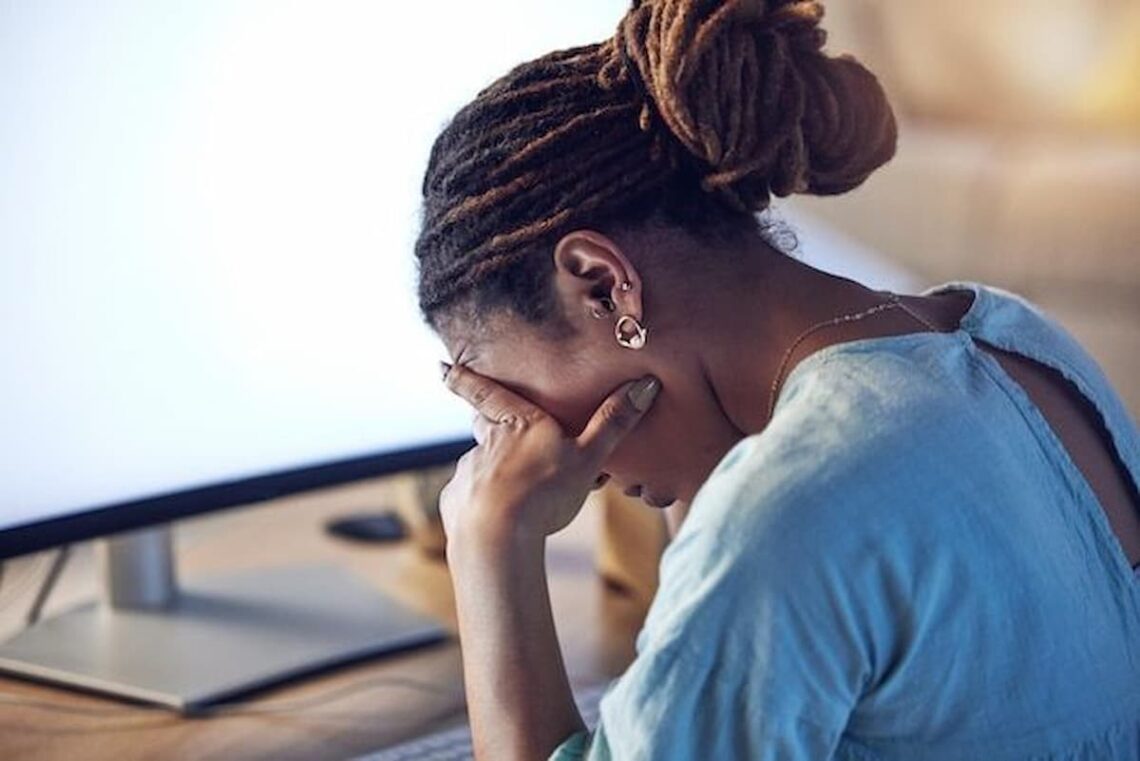A woman sitting at a desk with her head in her hand, looking stressed in front of a computer screen.