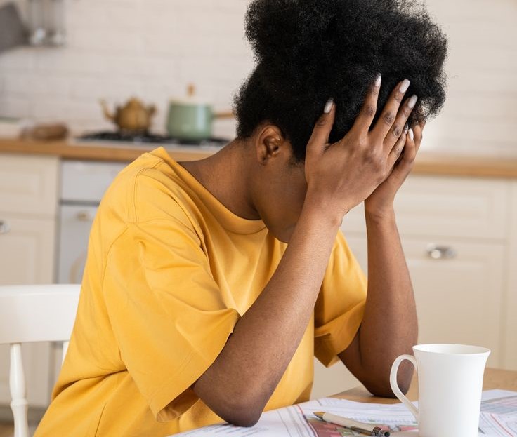 Black woman holding their head in their hands with office background, visually conveying emotional exhaustion