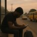 A young Nigerian man sits alone on the roadside beside a yellow danfo bus, staring thoughtfully into the distance.