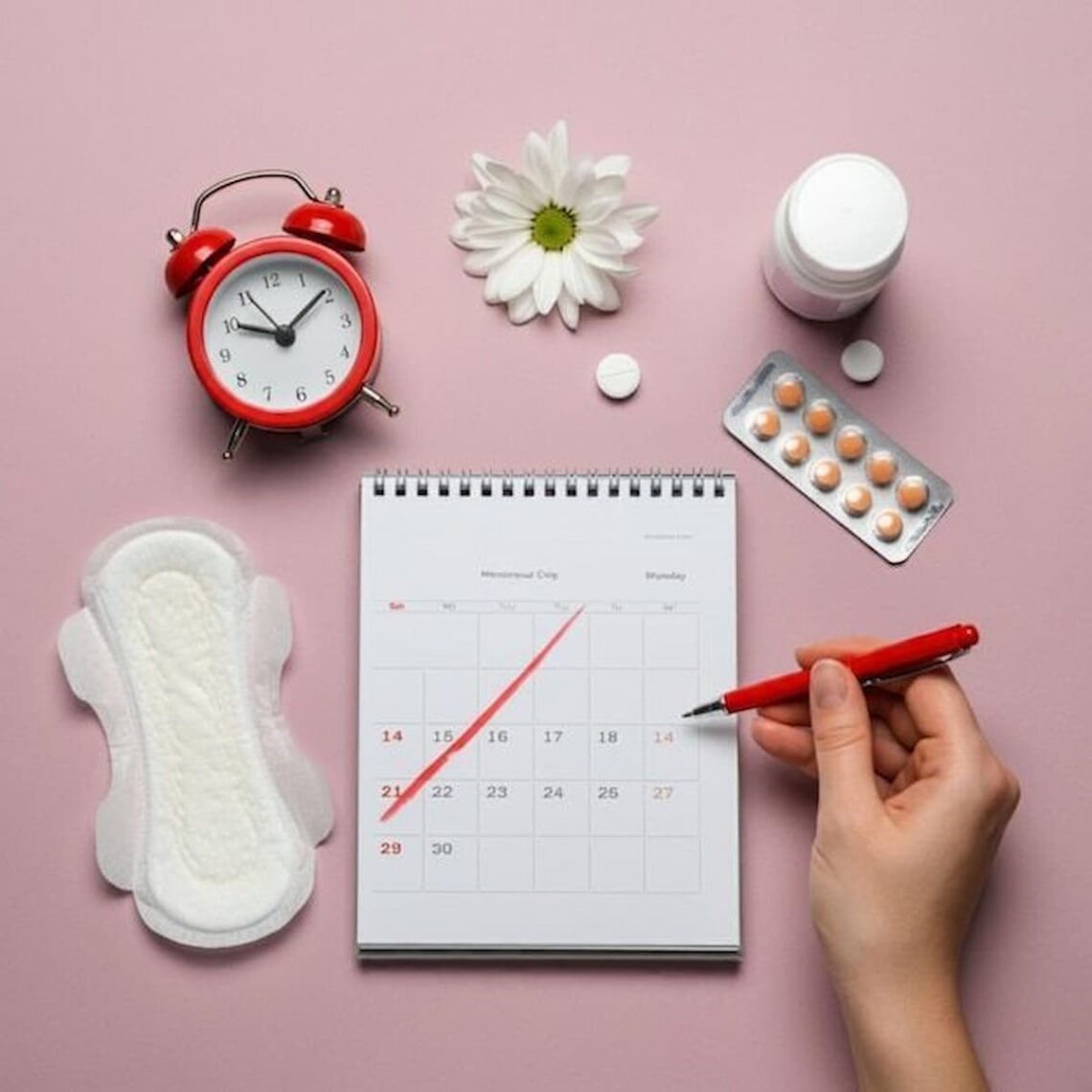 Flat lay on pink background showing a sanitary pad, period tracker calendar, red alarm clock, pain relief pills, daisy flower, and a hand marking dates with a red pen.