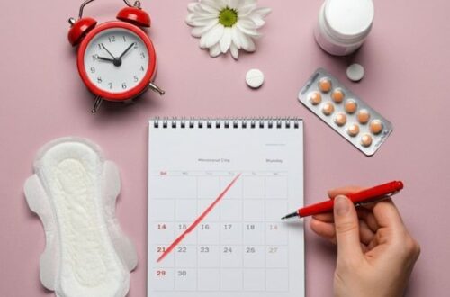 Flat lay on pink background showing a sanitary pad, period tracker calendar, red alarm clock, pain relief pills, daisy flower, and a hand marking dates with a red pen.